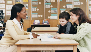 Teacher Sitting at a School Desk Showing a Book to a Parent and Her Son