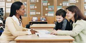 Teacher Sitting at a School Desk Showing a Book to a Parent and Her Son